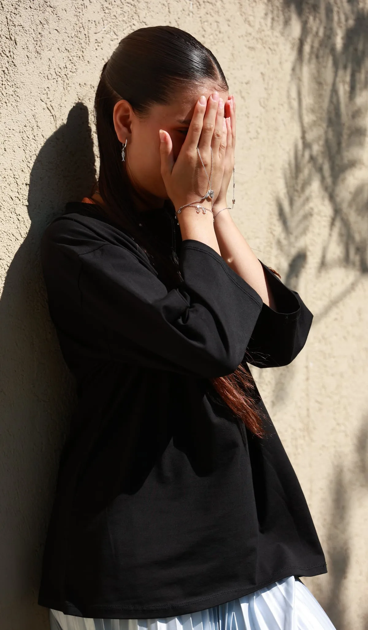 A young girl covering her face, leaning against a sunlit wall