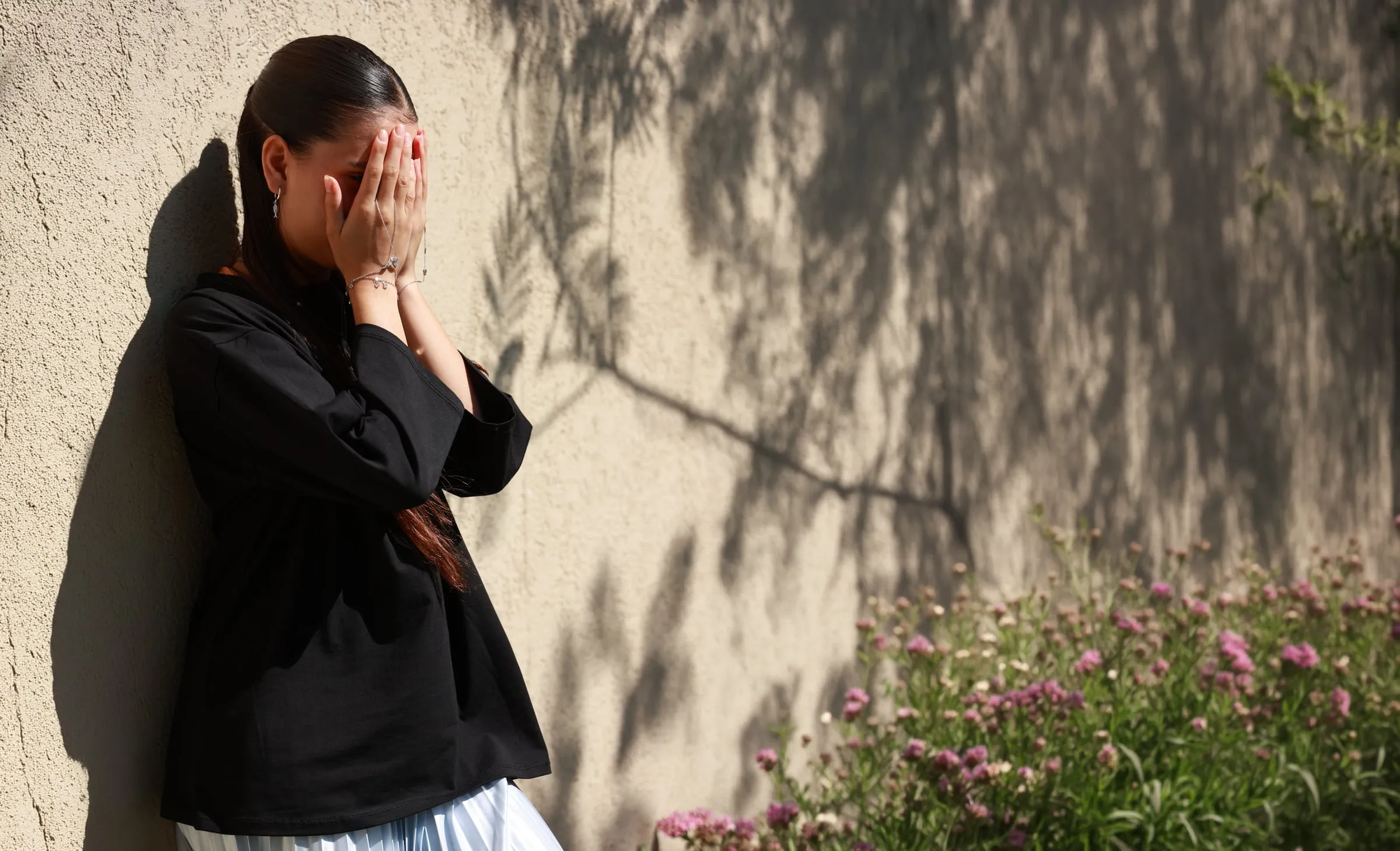 A young girl covering her face, leaning against a sunlit wall